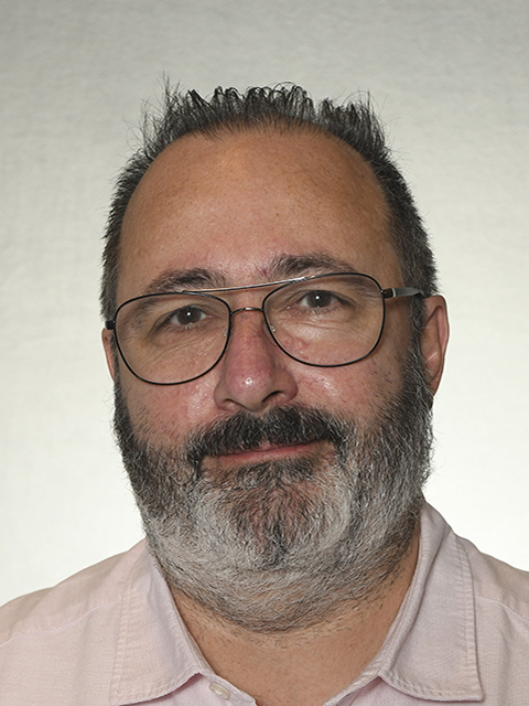 Person with short hair, glasses, and beard wearing a light pink shirt against a plain background
